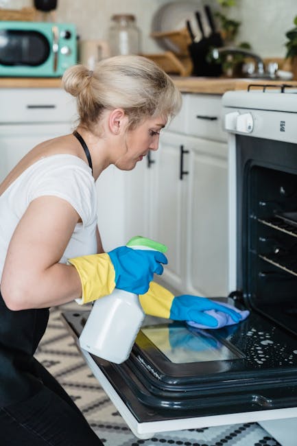 Close-up view of a black oven floor with soapy water droplets and foam, where a cleaning cloth with purple and pink hues is being used by a person wearing a yellow glove, demonstrating surface cleanin